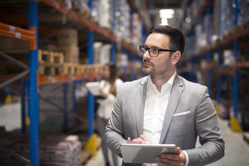 Warehouse manager reviewing IP6 on a tablet between the shelves, with a colleague in the background